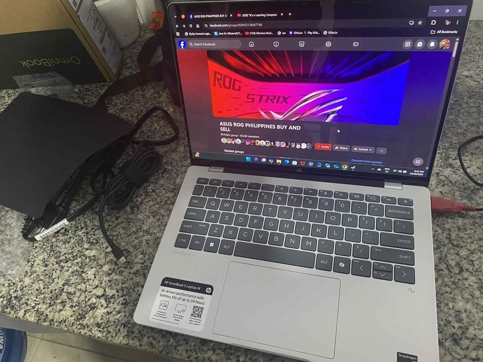 Hands-on photo of the HP OmniBook 5 OLED Snapdragon X laptop, showing the keyboard, trackpad, and included power adapter